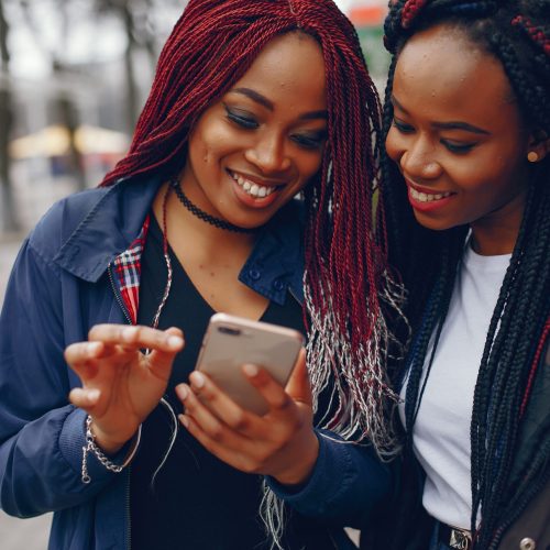 two beautiful and stylish dark-skinned girls with long hair standing in a autumn city near purple wall and using the phone